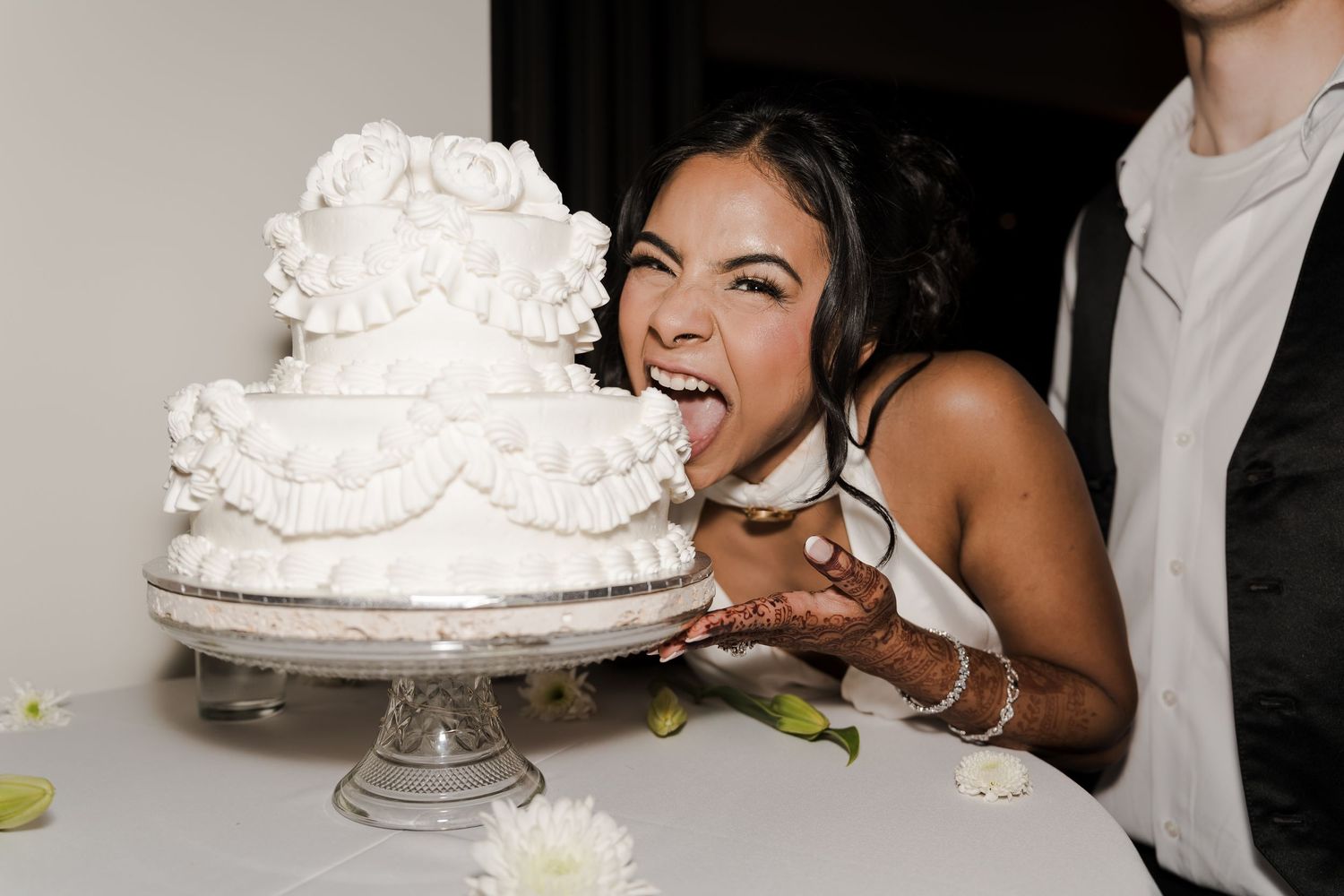 Bride joyfully biting into a white tiered wedding cake with ruffled decorations at a wedding reception.