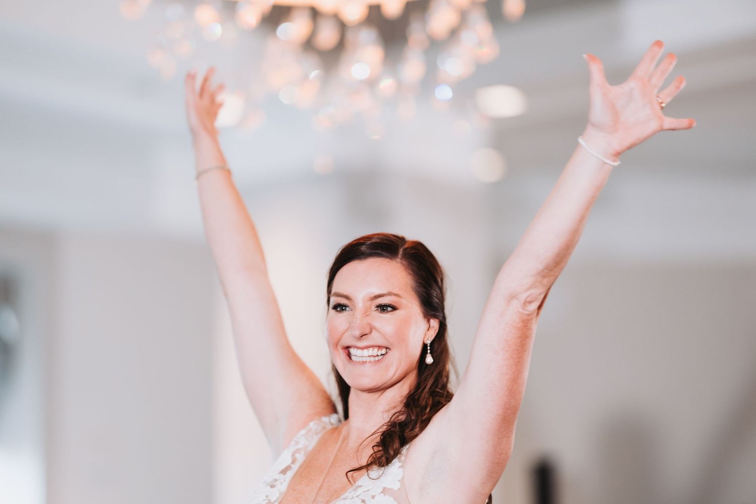 A bride in a white wedding dress raises her arms joyfully during an outdoor celebratory moment.