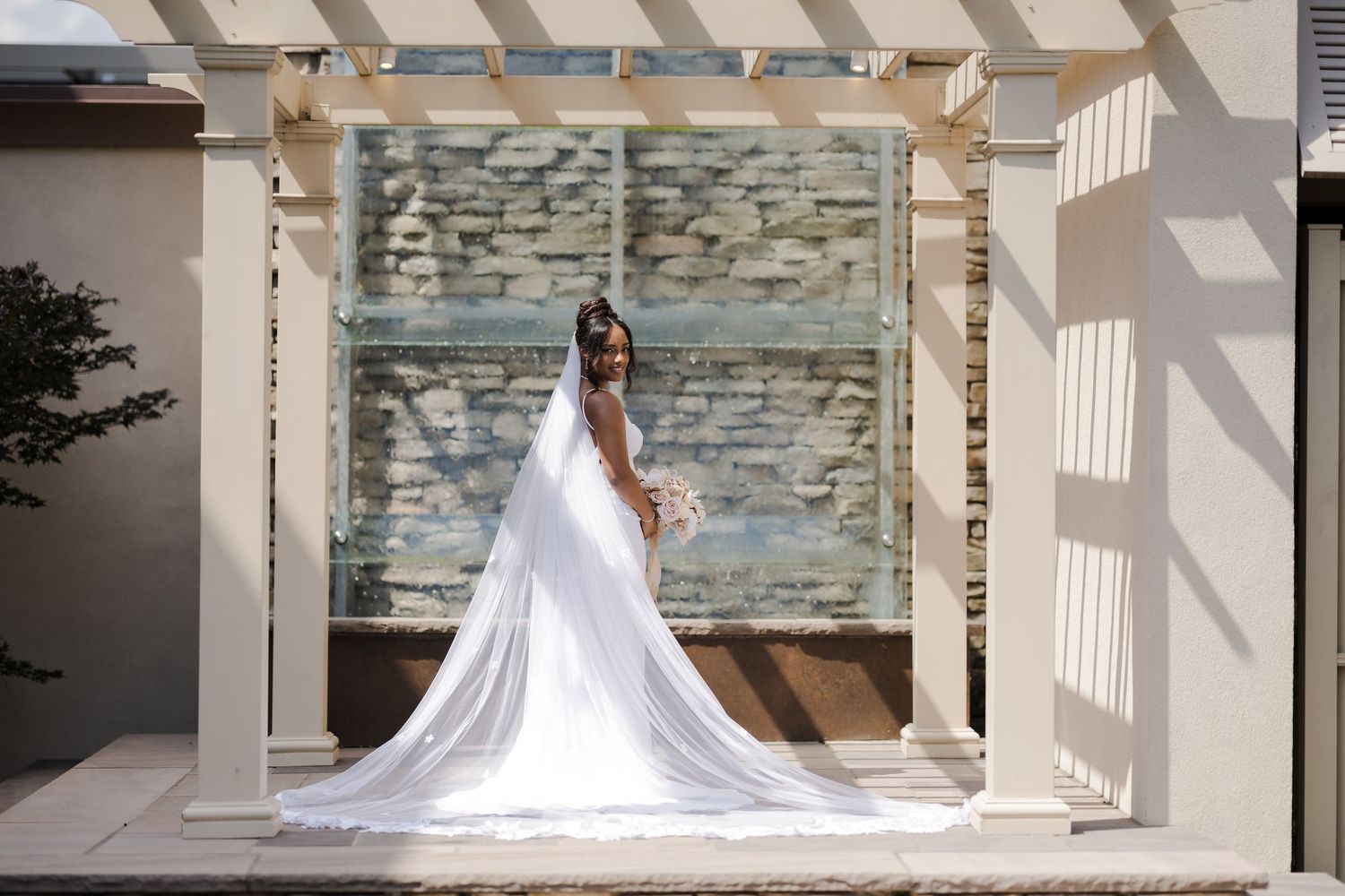 A flowing white wedding gown with a long train stands under an elegant white pergola against a stone wall backdrop.