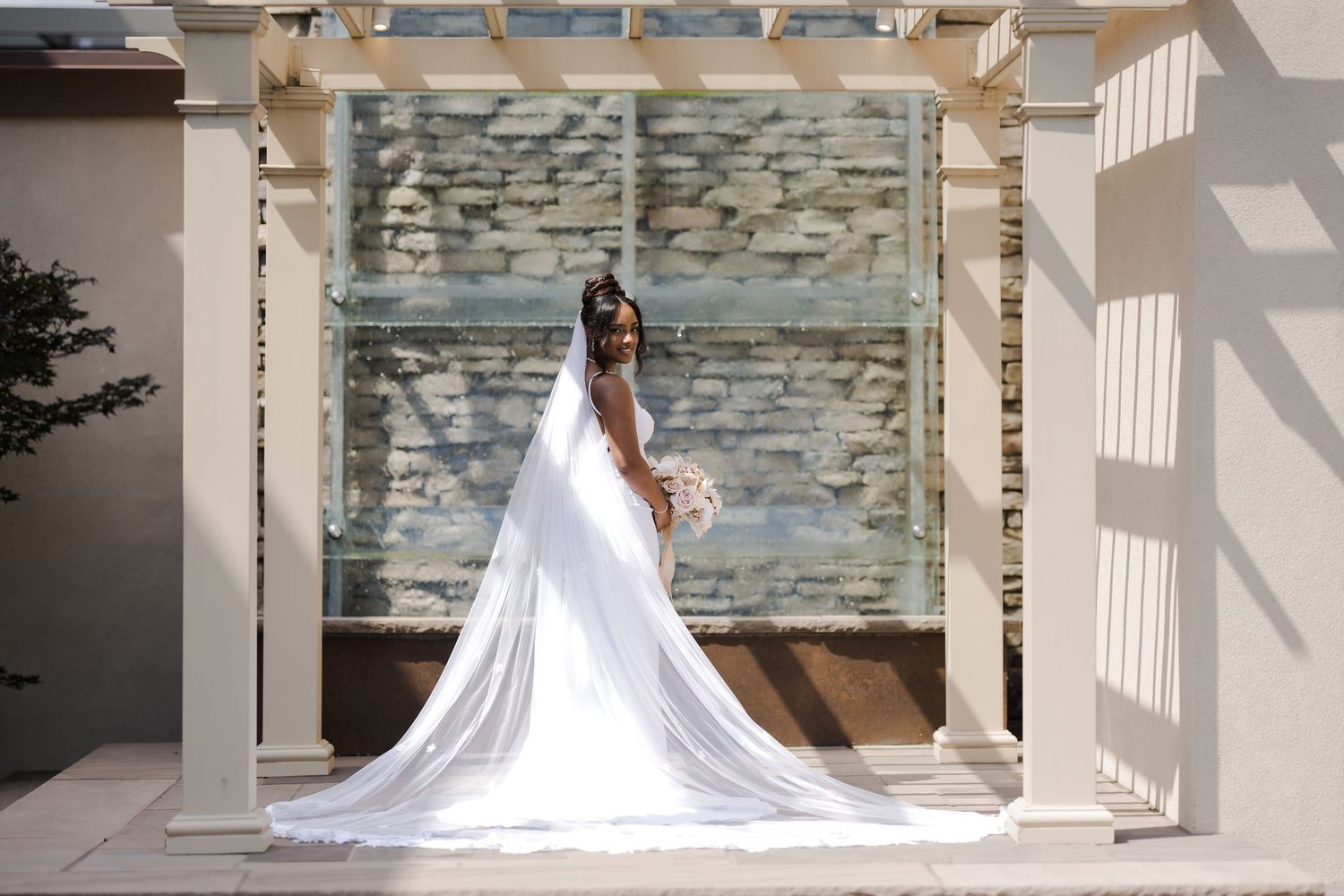 A bride in a flowing white wedding gown with a dramatic long train poses in a sunlit hallway with stone walls.