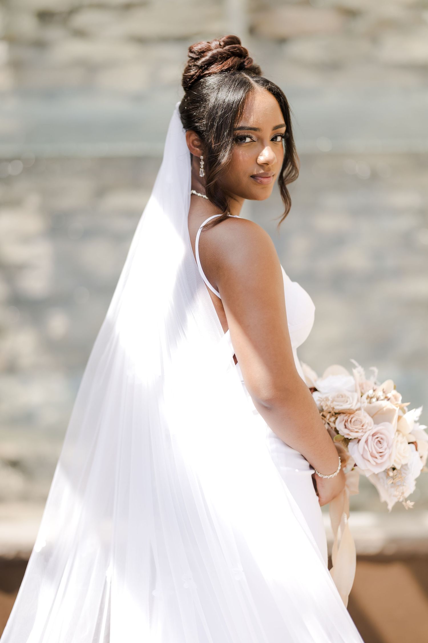 A bride in an elegant white wedding dress with a flowing veil holds a soft pink bouquet against a stone background.