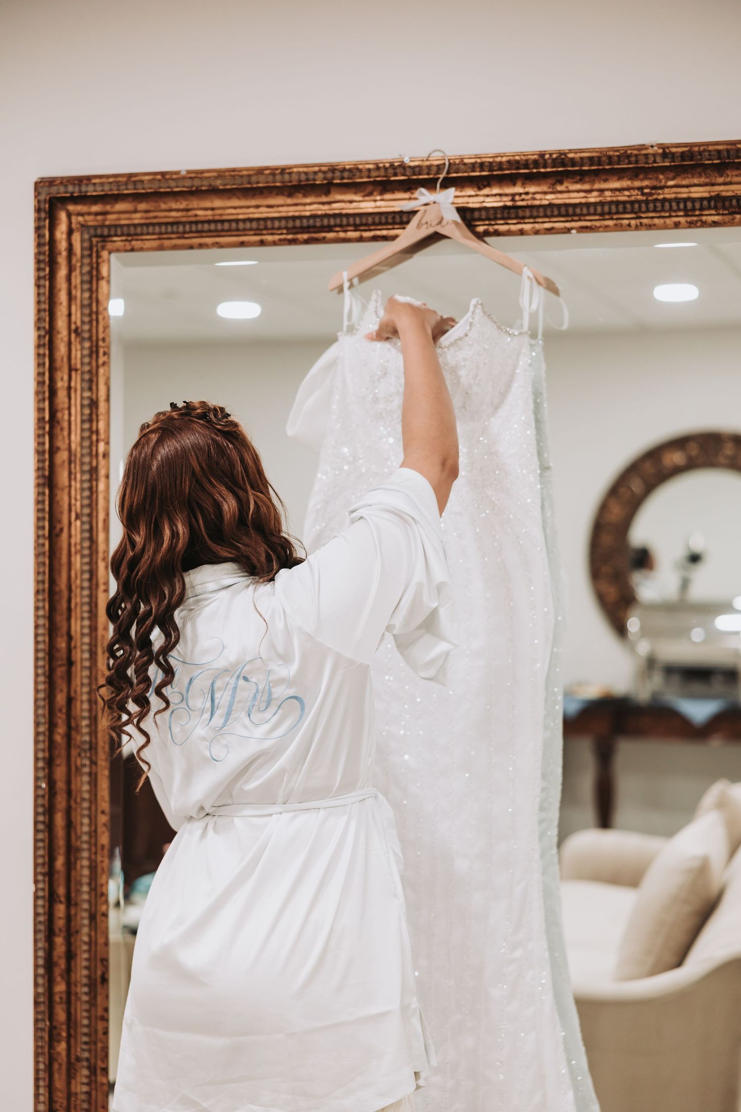 Person in white robe reaching up to wedding dress hanging on ornate gold-framed mirror.