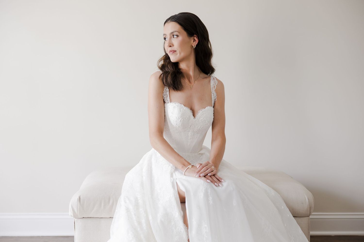 A person in a white wedding dress sits gracefully on a light-colored couch against a neutral wall.