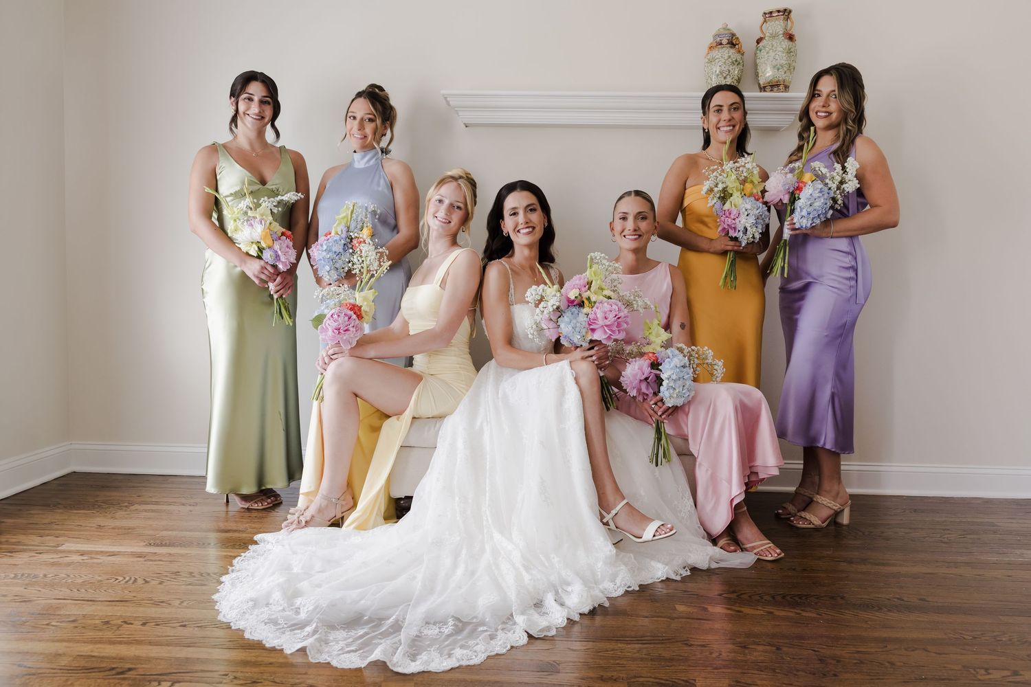 Wedding party wearing pastel slip dresses in yellow, purple, and sage poses with bride in white lace gown with floral bouquets.