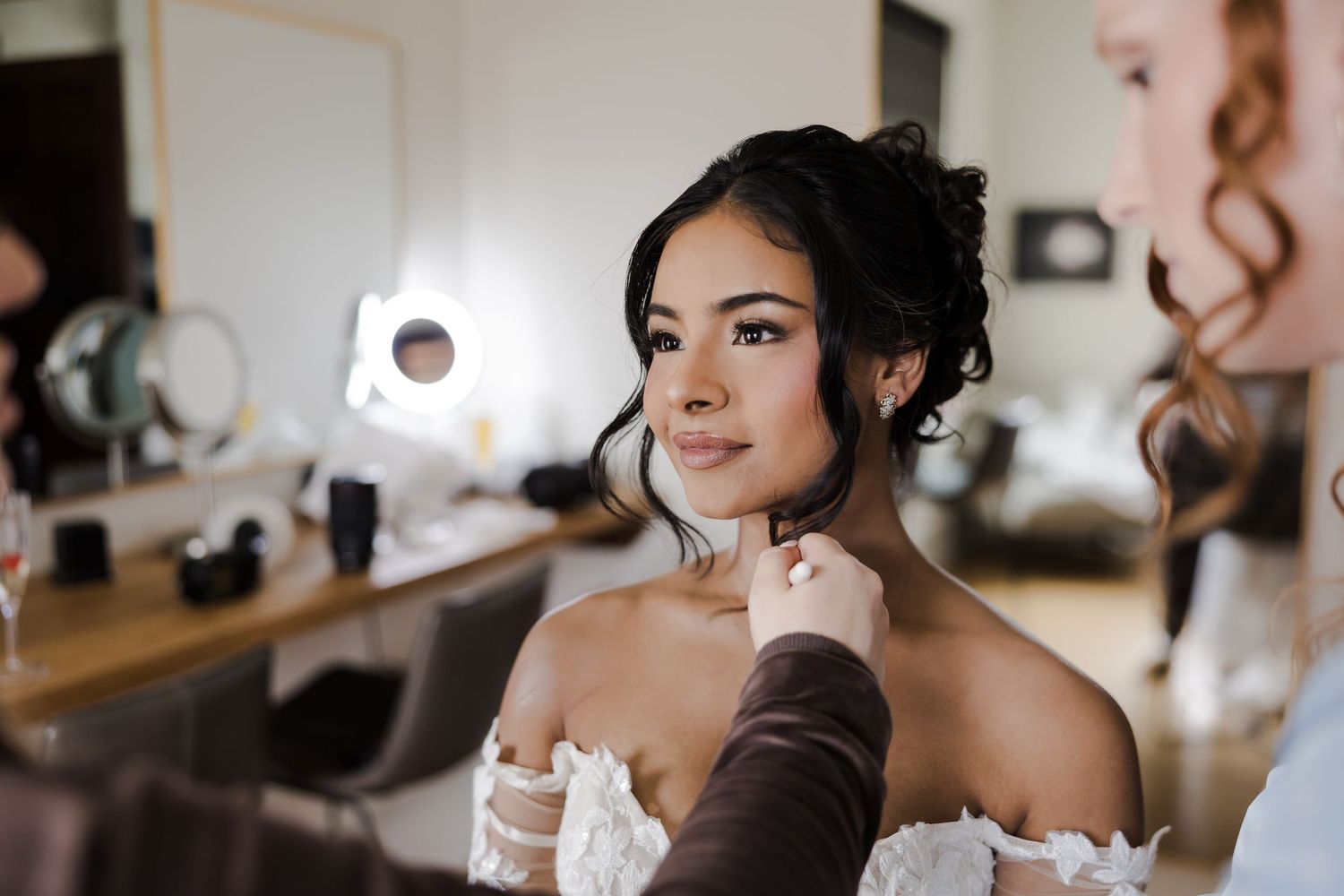 Bride getting jewelry put on while sitting in front of a vanity mirror during wedding preparations.