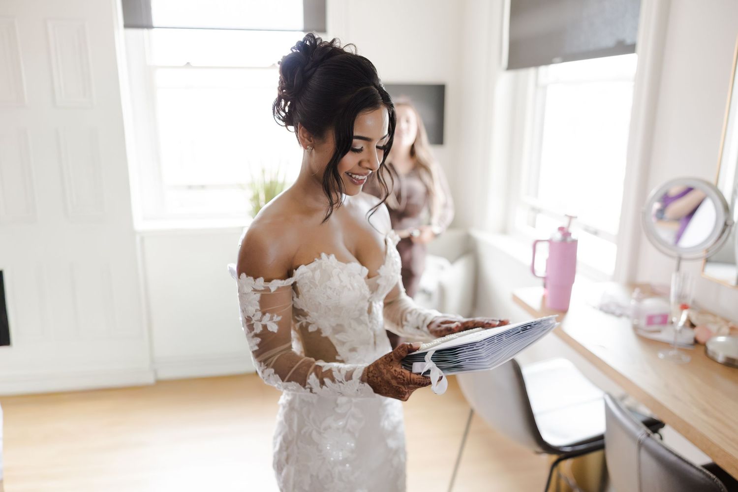 Smiling bride in lace off-shoulder wedding dress reading a card while getting ready in a bright dressing room.
