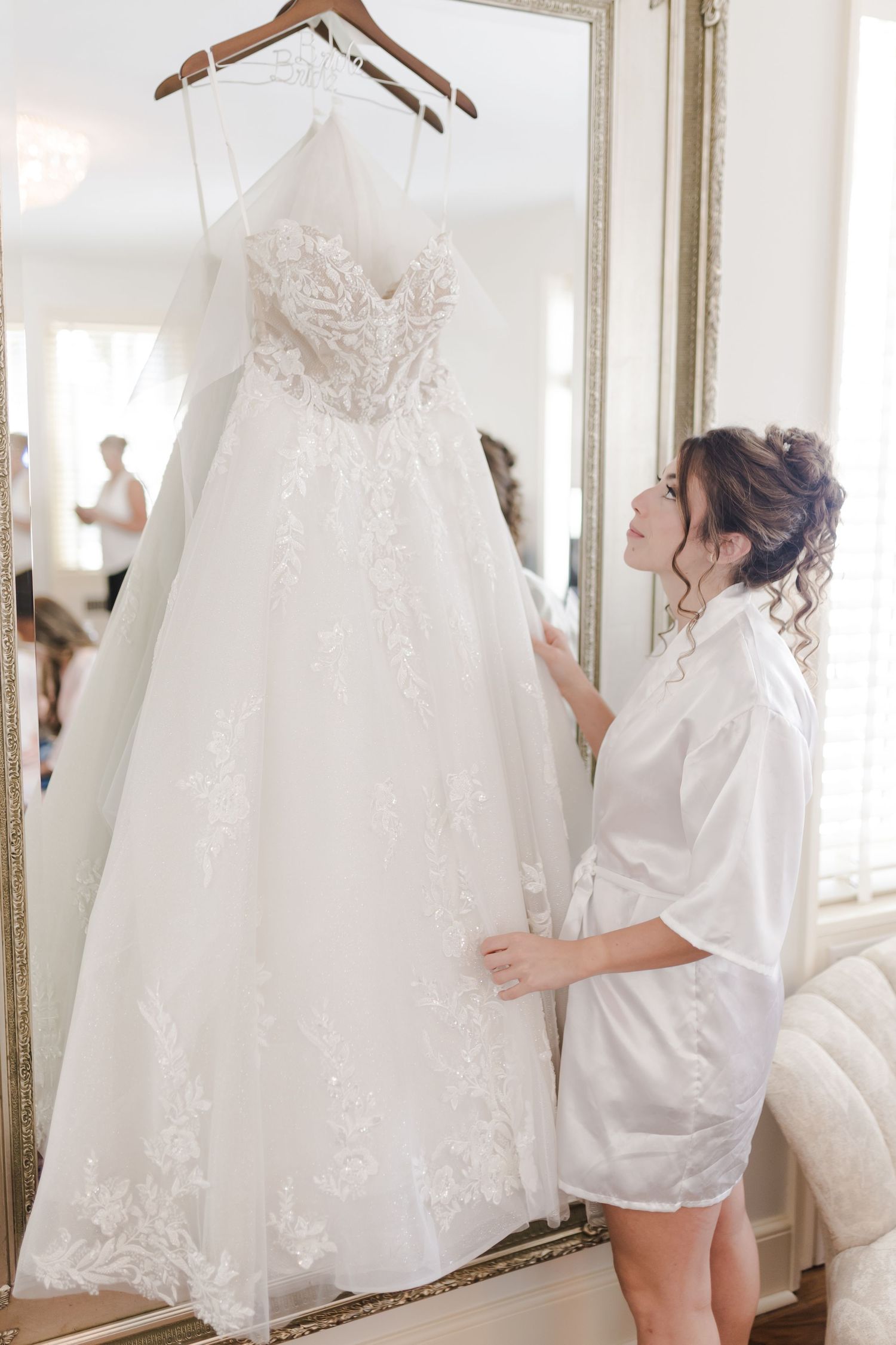 Wedding dress with delicate lace details hangs on display as someone in a white robe admires it in a mirror.