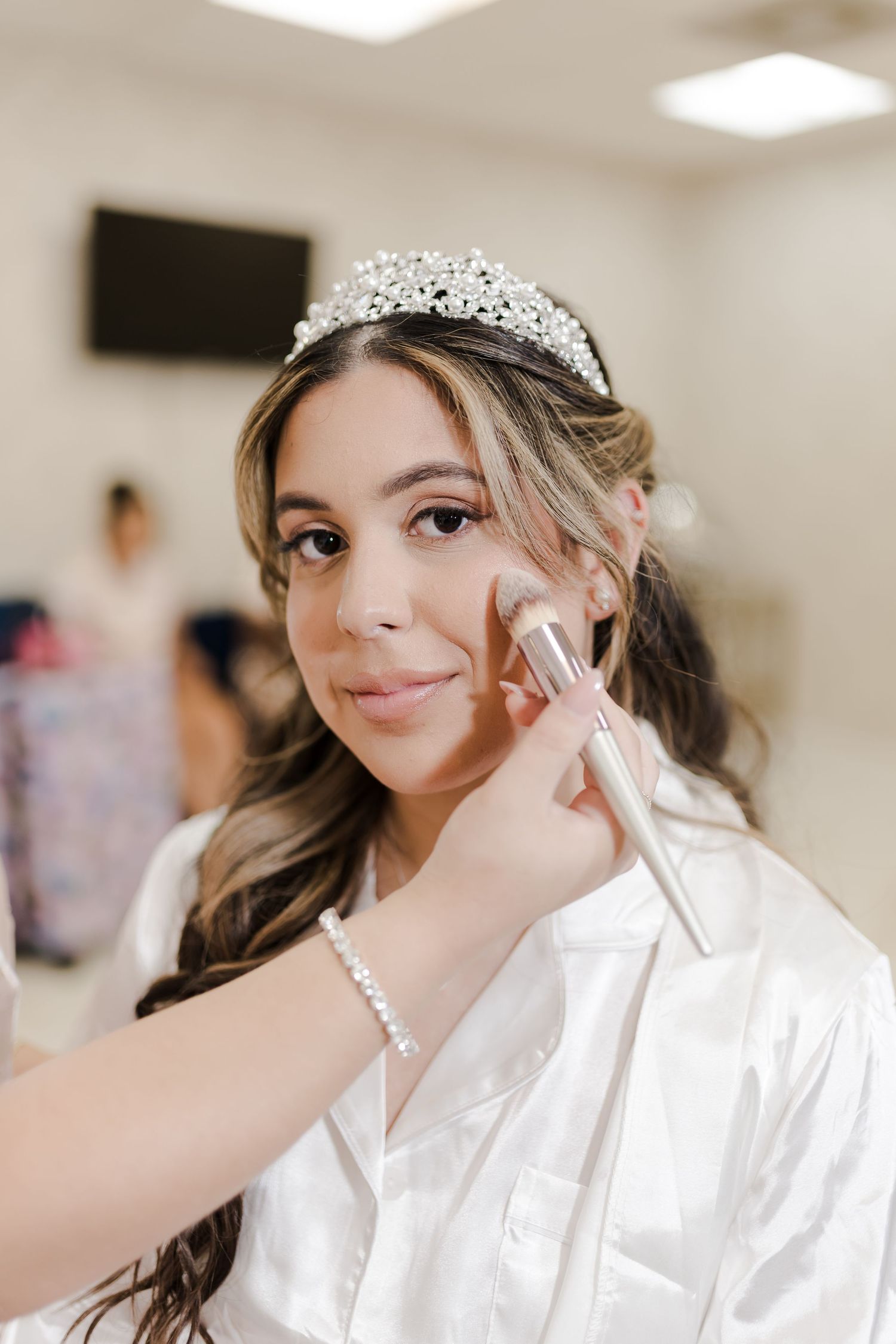 Someone wearing a bridal pearl tiara and white robe getting makeup applied while preparing for a wedding ceremony.
