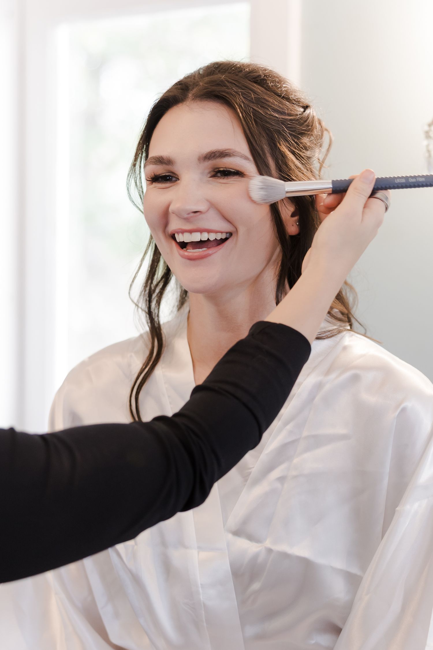 Makeup artist applies cosmetics with brush to smiling client in white robe during professional beauty session.