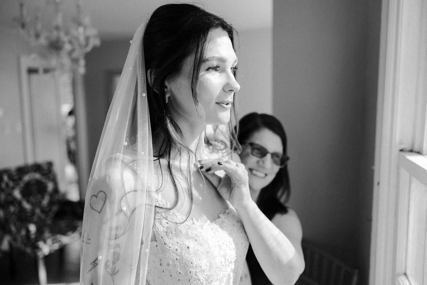 A bride in a white wedding dress and veil stands by a window during wedding preparations.