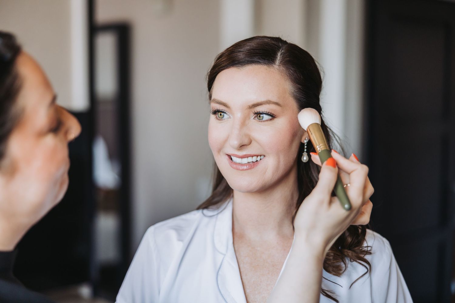Getting ready for a wedding day, a makeup artist applies cosmetics to a smiling person wearing white.