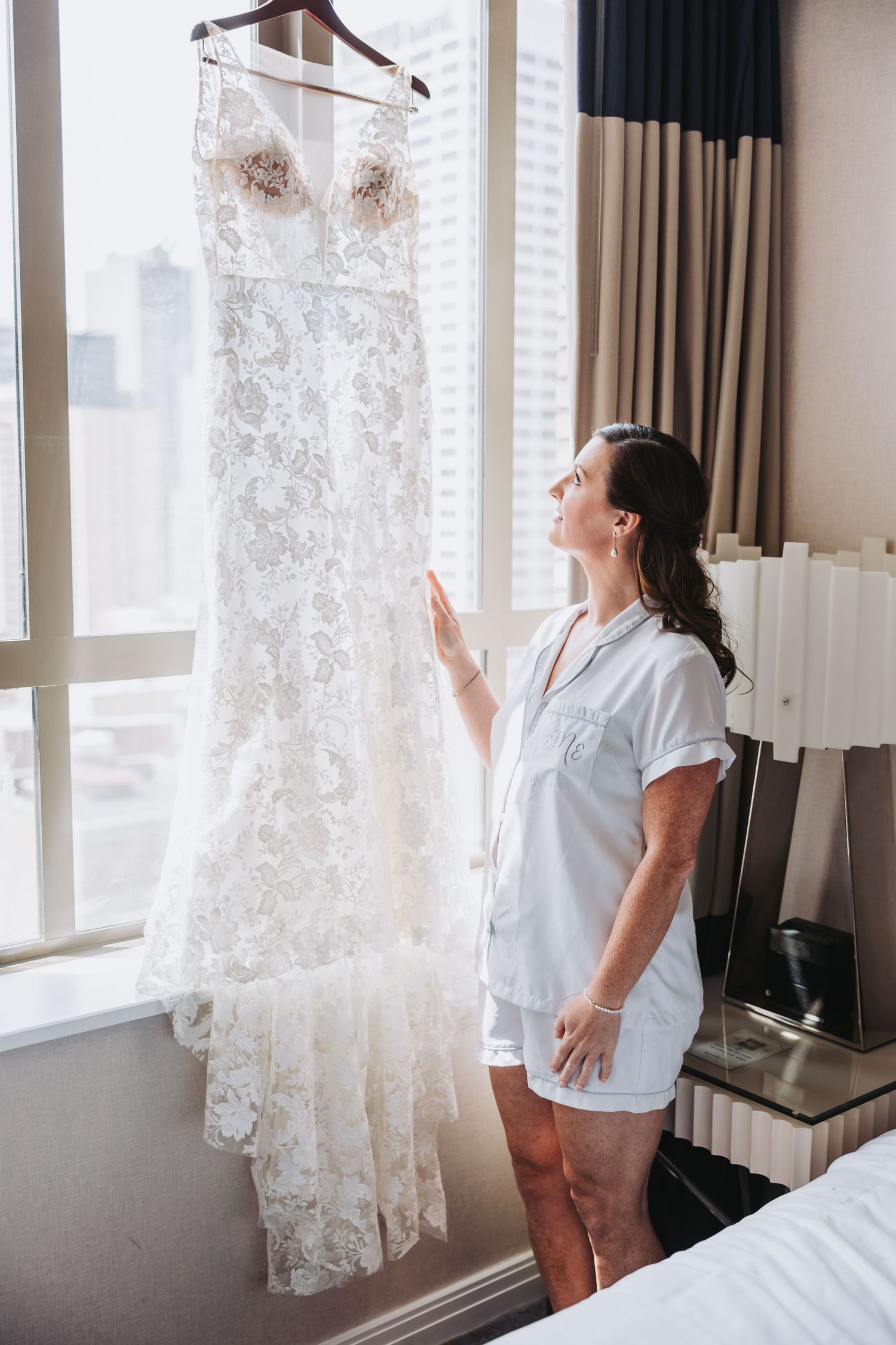 A white lace wedding dress hangs by a window while someone in a robe admires it in a hotel room.