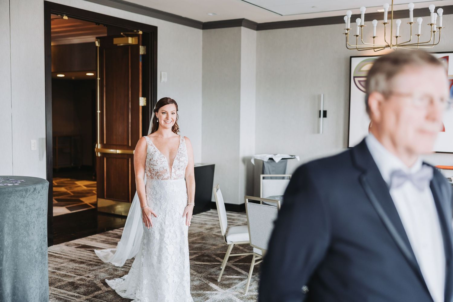 A person in an elegant white wedding gown stands in a hotel venue while someone in formal attire walks by.