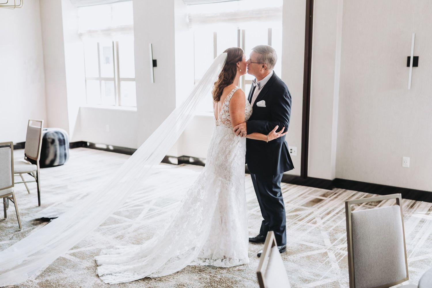 A bride and groom share an intimate moment while dancing in a bright, white room with large windows.