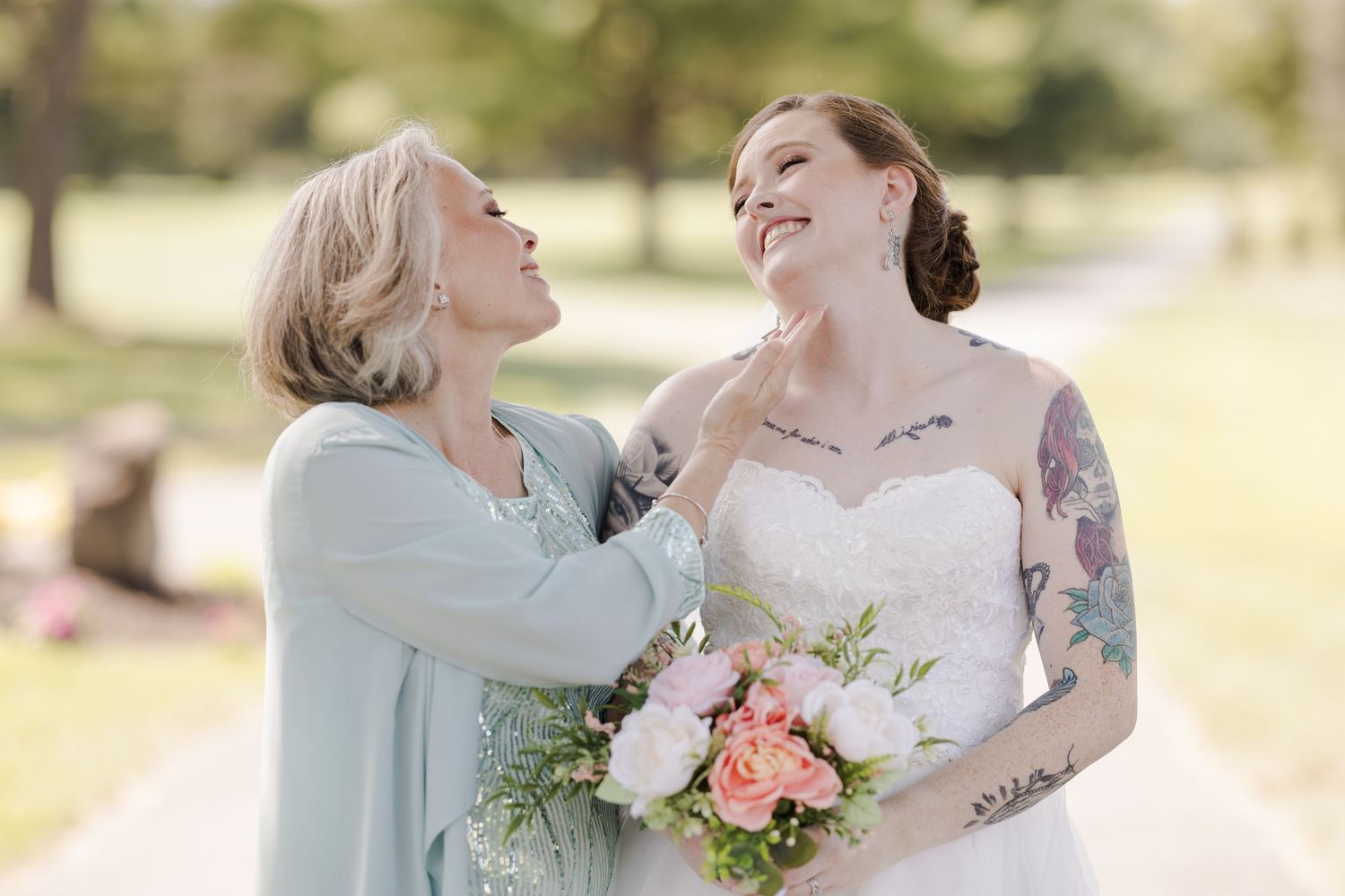 Bride in white gown holding pink bouquet smiles as older woman in blue adjusts her necklace outdoors.