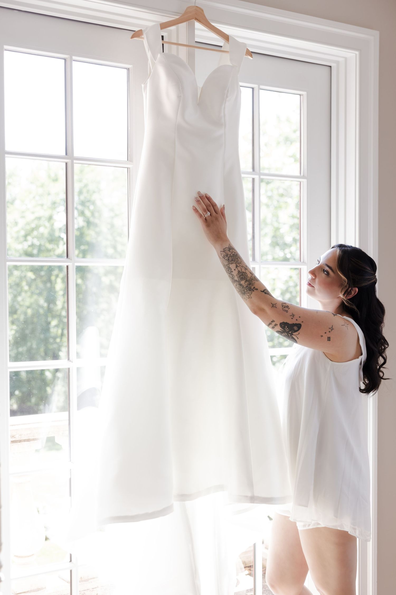 Tattooed bride in white robe admiring her wedding dress hanging by a bright window.