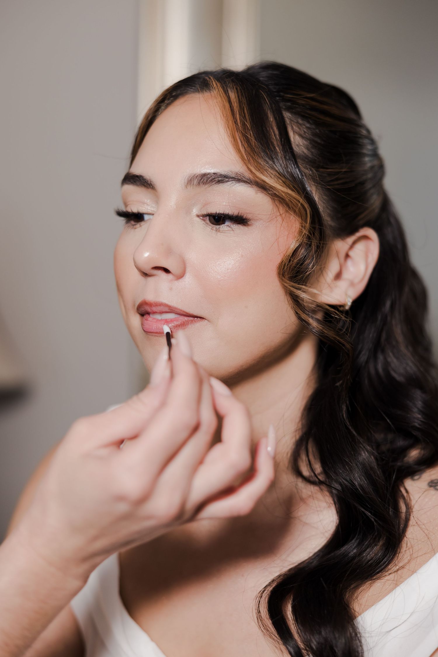 Bride applying nude lipstick while getting ready, wearing white dress with elegant wavy dark hair.
