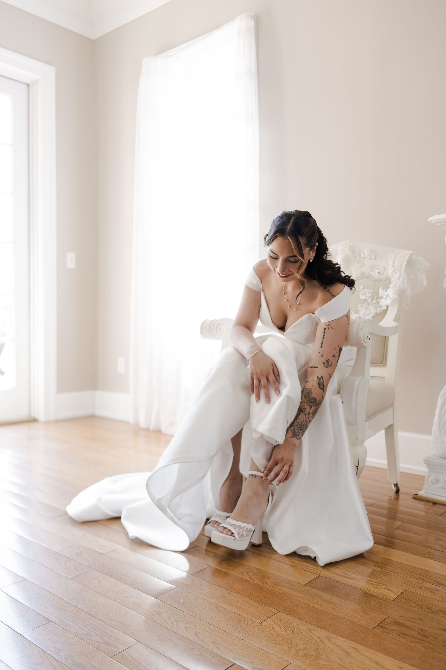 Bride in white off-shoulder gown sits on chair putting on heels, tattoos visible, natural light room.