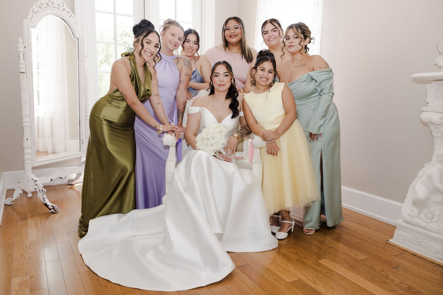 Bride in white gown poses with bridesmaids in colorful satin dresses inside an elegant bright room.