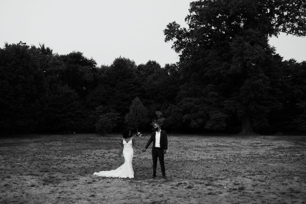 A couple stands together in an open field at dusk with trees silhouetted in the background.