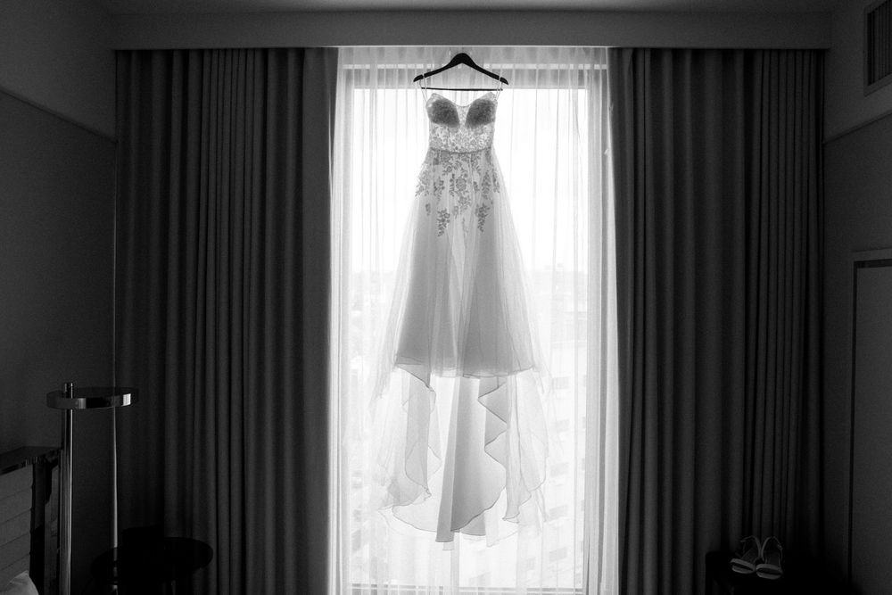Elegant white wedding dress hangs by a bright window creating a dreamy silhouette against light curtains.