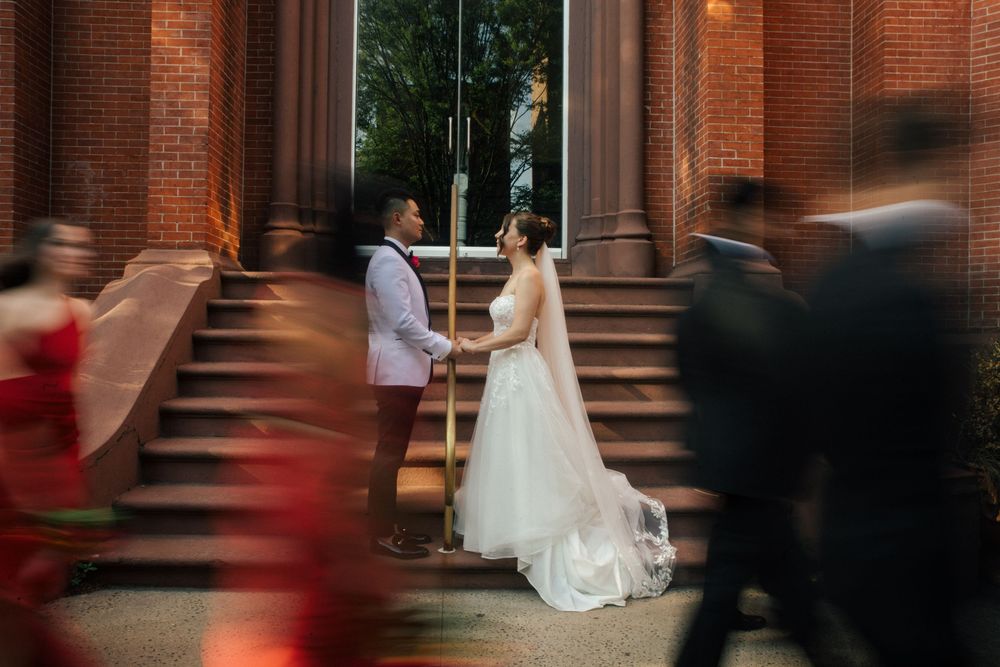 Motion-blurred scene in a grand brick building showing wedding guests moving past a seated figure in white.