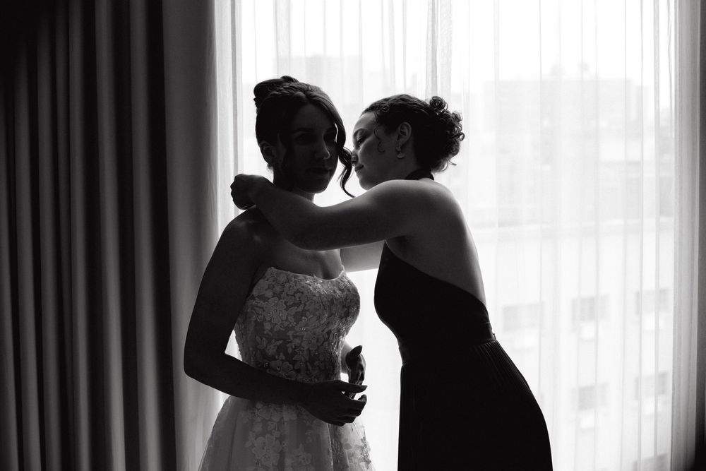 A tender silhouette moment between a bride in a wedding dress and woman assisting her by a bright window.