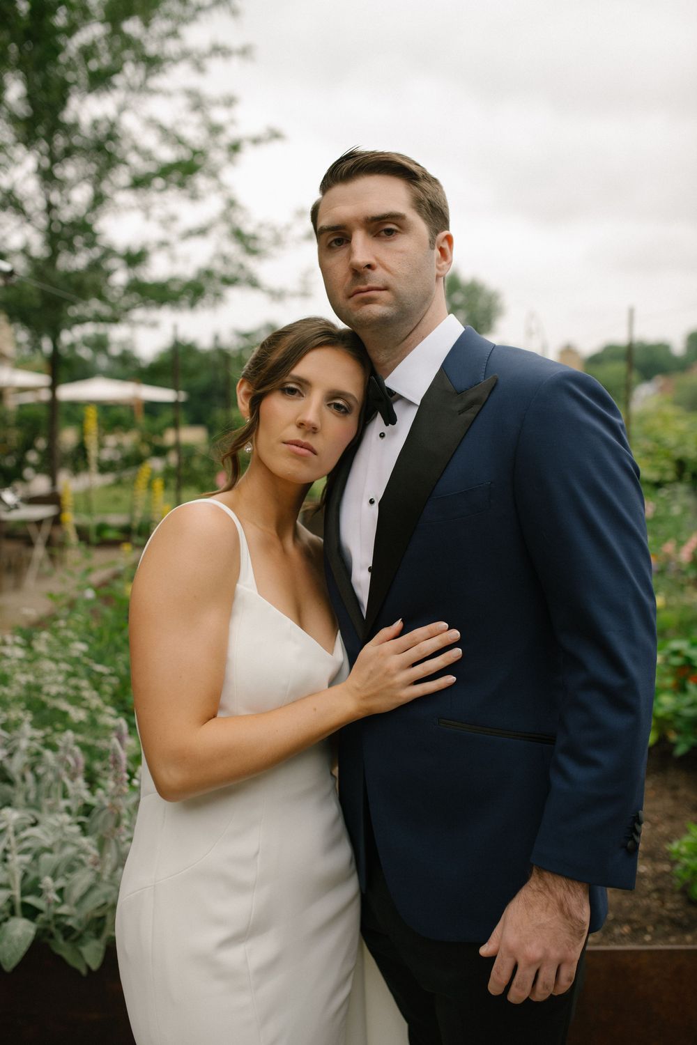 A couple share intimate wedding portraits in a garden with lush greenery and overcast skies.