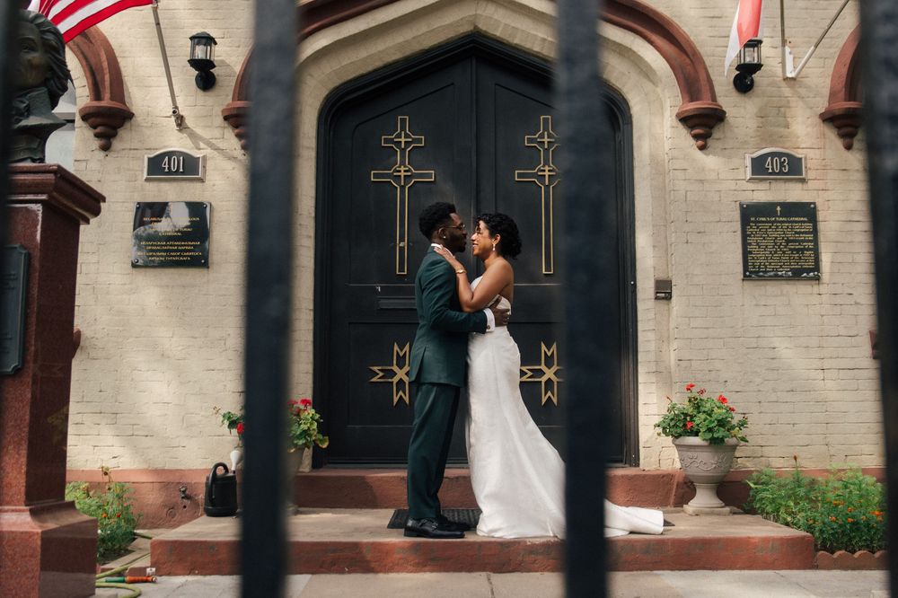 Wedding couple in elegant attire stands in front of a church entrance with American flag and crosses in view.