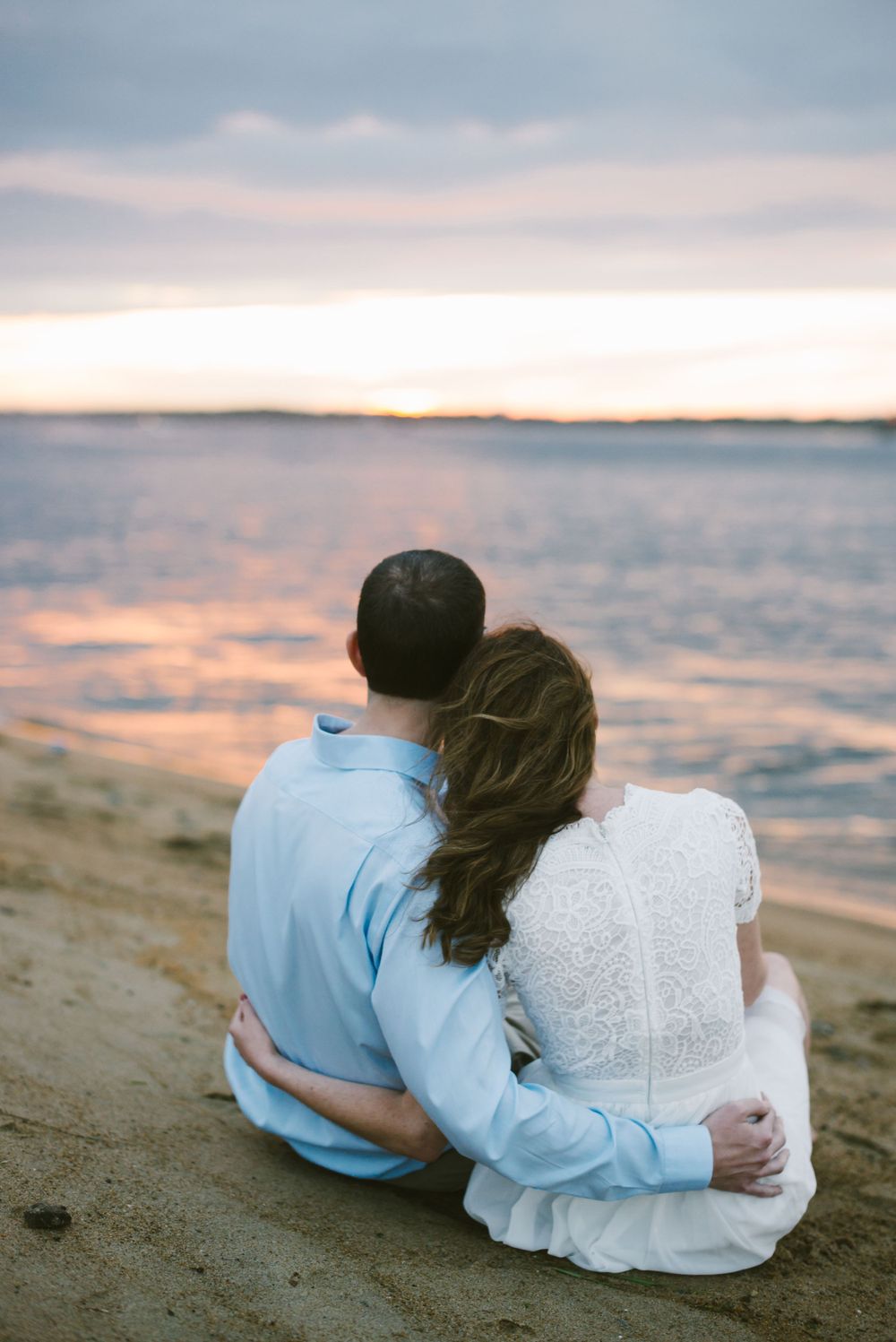 Couple sits close together on beach watching colorful sunset over calm waters.
