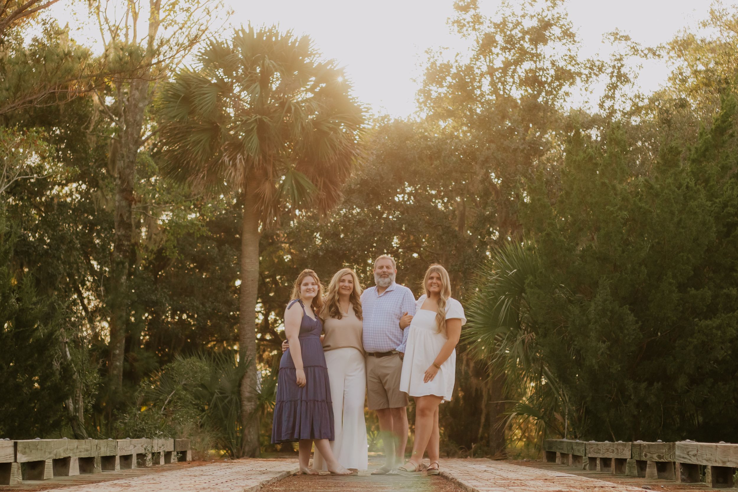 Family portrait on wooden bridge during golden hour with sunlit trees in background.