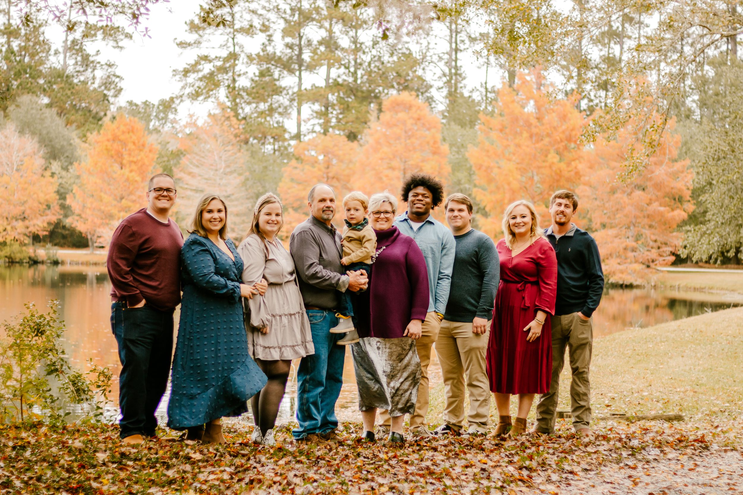 Group portrait of extended family in fall clothing against autumn foliage and lake background.