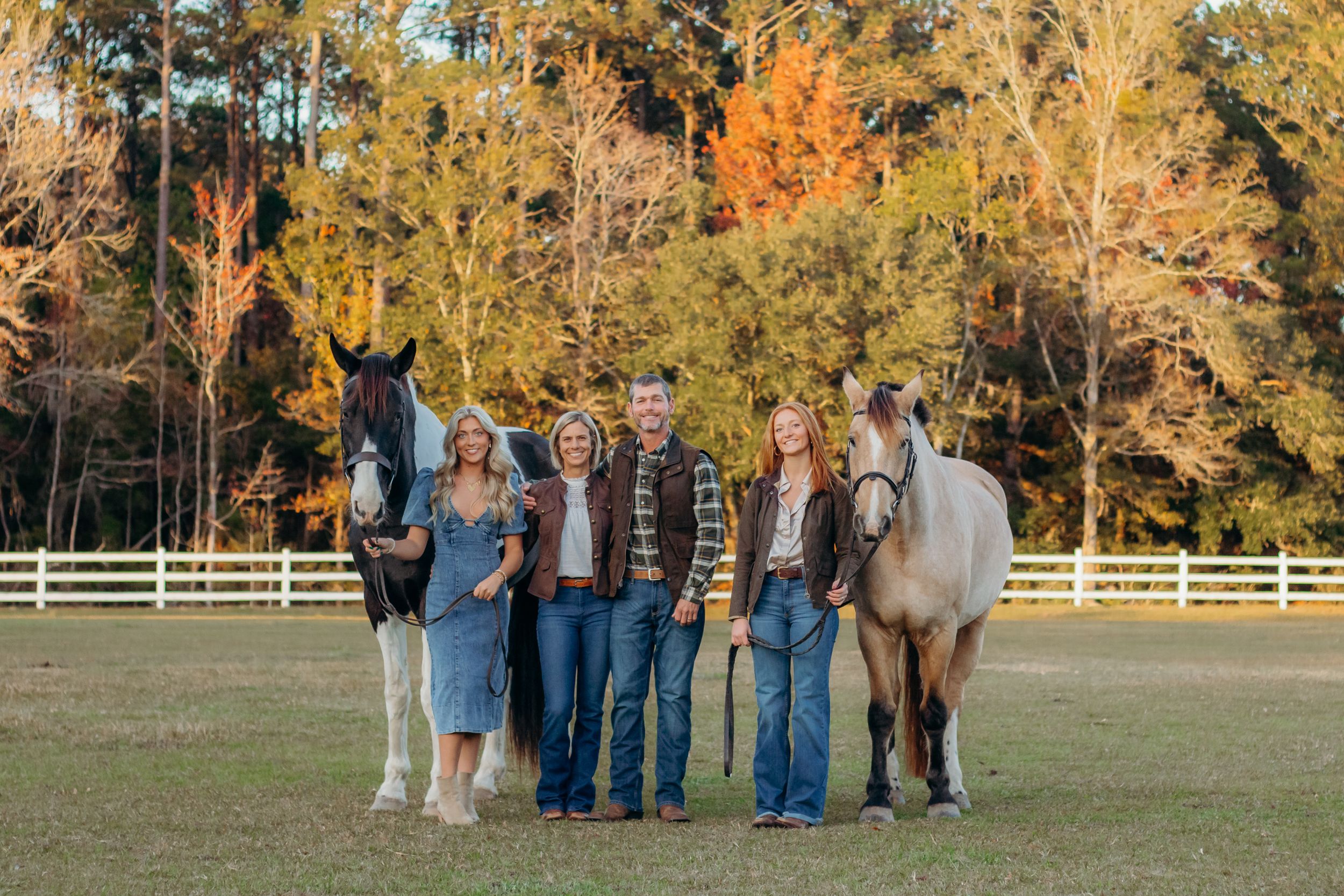 Family group portrait with horses in a fall setting with white fence and autumn foliage.