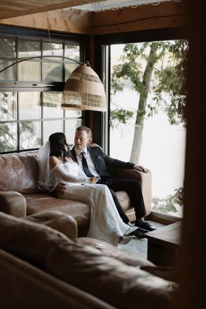 A couple lounges on a leather couch in a rustic room with woven pendant lamps and large windows.
