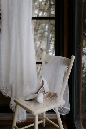 Elegant white wedding dress hanging by window with rustic wooden chairs in foreground.