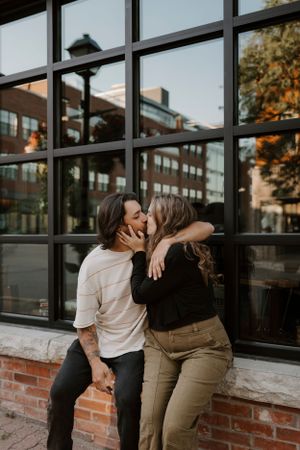 Two people embrace while sitting outside a commercial building with large windows.