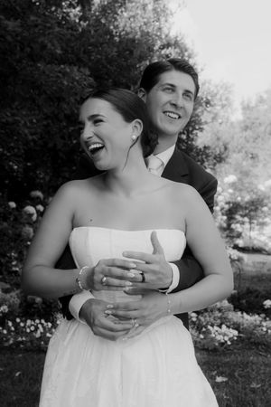 Wedding party members in white dresses share joyful laughter in a garden setting.