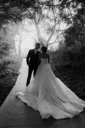 Black and white photo of couple walking on wooden path with dramatic train of wedding dress.