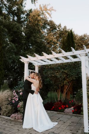 Wedding couple poses beneath a white pergola in a garden setting with vibrant red flowers.