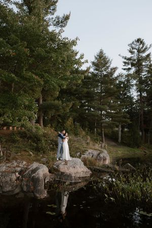 Wedding couple posing on scenic rocky cliff overlooking serene lake and pine forest.