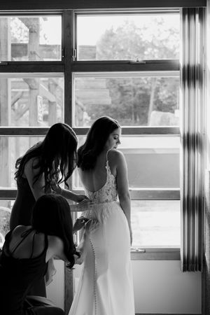 Wedding preparation scene in silhouette showing dress adjustments near a bright window.