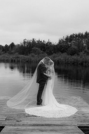 Black and white photo of newlyweds embracing on a dock overlooking a serene lake.