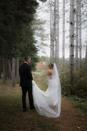 Bride's flowing veil trails behind as couple walks through misty forest path.