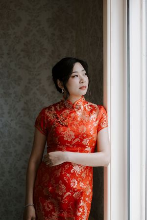 Traditional red Chinese qipao dress with gold floral embroidery photographed near a window in dramatic lighting.