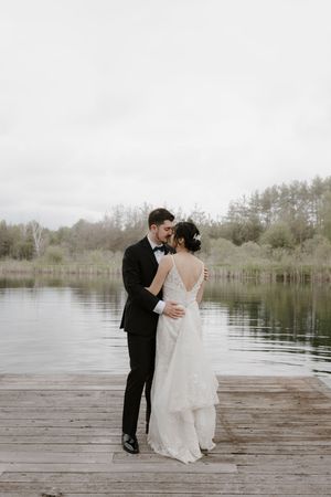 Couple in wedding attire dances on wooden dock with lake backdrop.