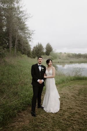 Couple in wedding attire poses together on grassy lakeside terrain.