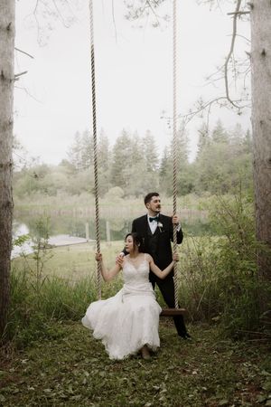 Newlyweds enjoy playful moments on a rustic rope swing surrounded by tall trees.