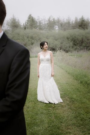 A bride in a white lace gown sits gracefully in a meadow near evergreen trees.
