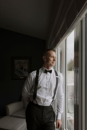 Groom in formal attire gazes thoughtfully out bright windows.
