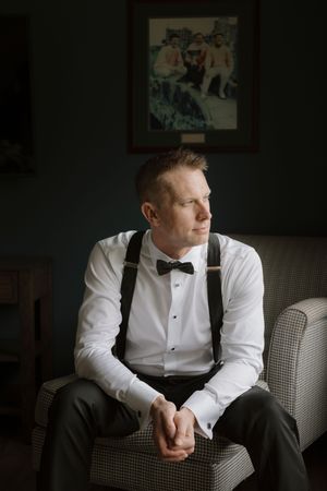 Groom in suspenders and dress shirt sits pensively in moody indoor setting.